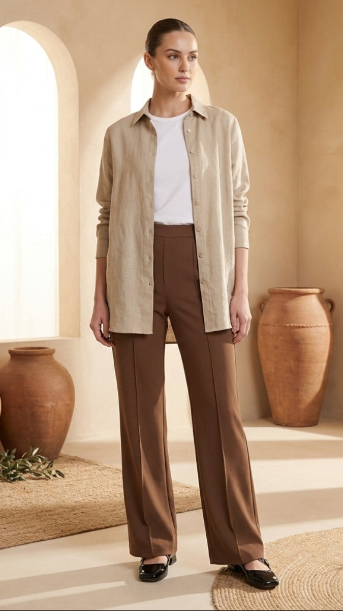 Woman in beige shirt and brown pants standing in a sunlit room with large pots and plants.