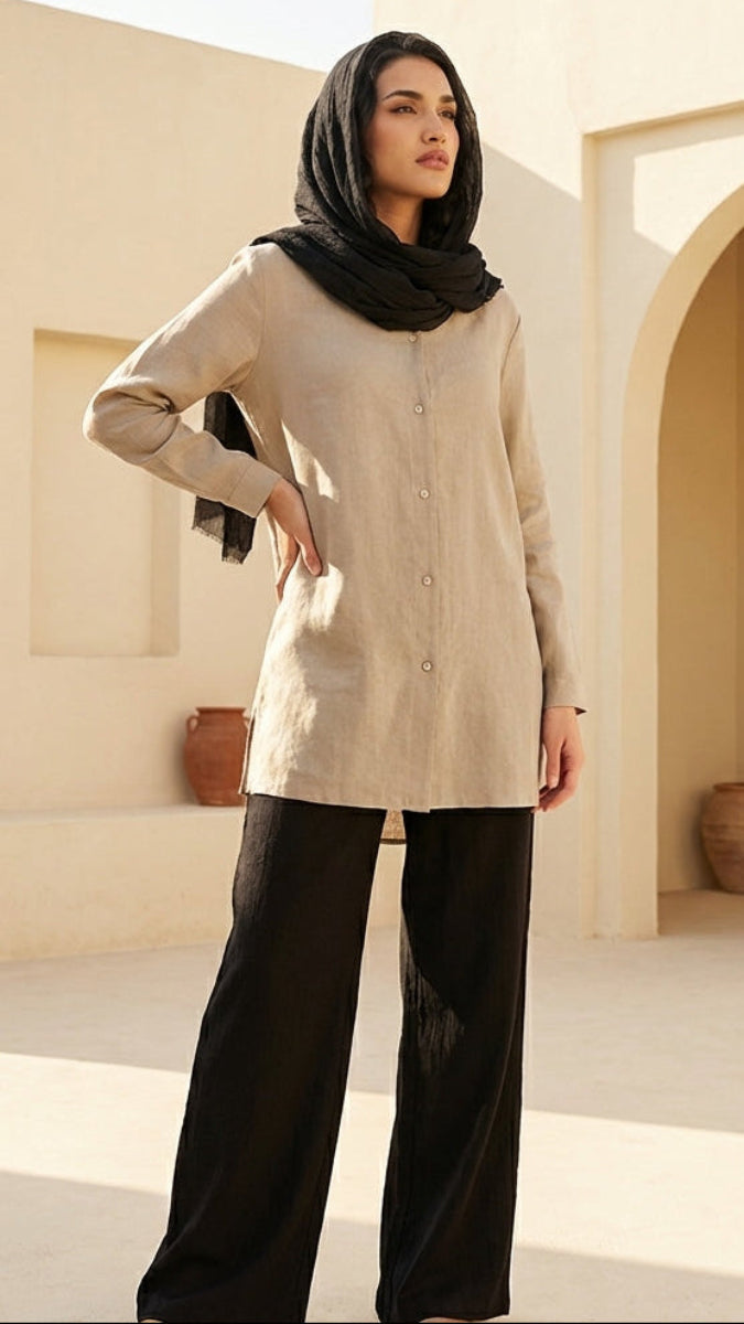 Woman in beige top and black pants standing in a sunlit outdoor setting with arches and pots.