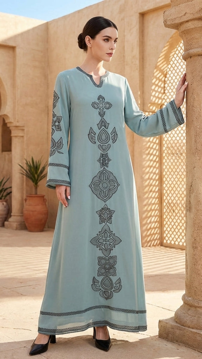 Woman in a long, light blue dress with intricate patterns standing in a desert setting with architectural elements.