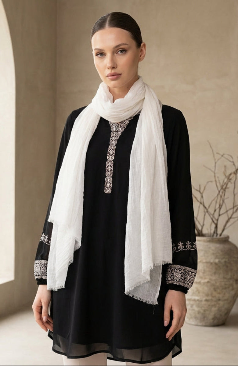 Woman wearing a black dress with white embroidery and a white scarf in a minimalistic setting.