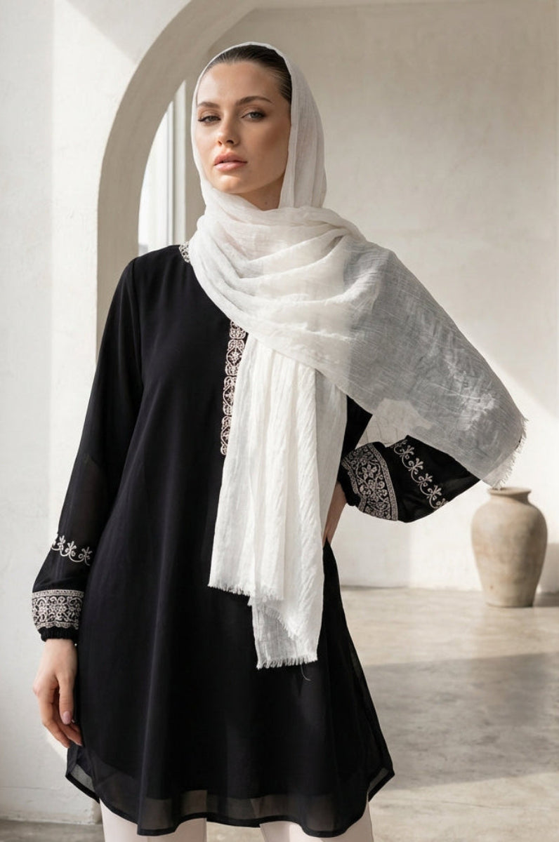 Woman wearing a black outfit with white embroidery and a white scarf in a bright room.