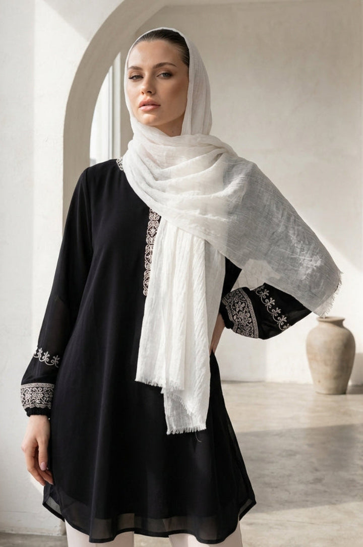 Woman wearing a black outfit with white embroidery and a white scarf in a bright room.