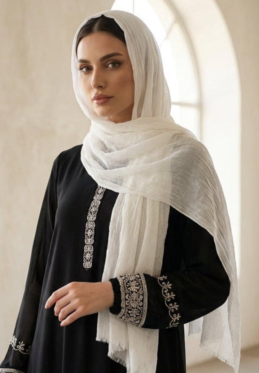 Woman wearing a black dress with white embroidery and a white hijab in a bright room.