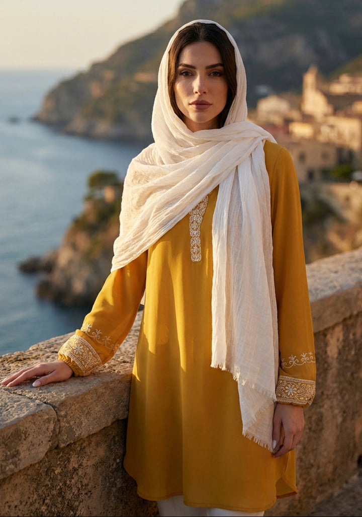 Woman in a yellow dress and white hijab standing by a scenic coastal wall.