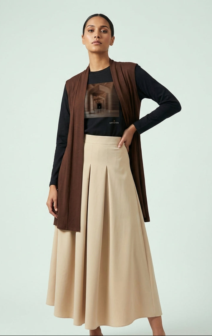 Woman wearing a brown vest, black long-sleeve shirt, and beige skirt on a light gray background