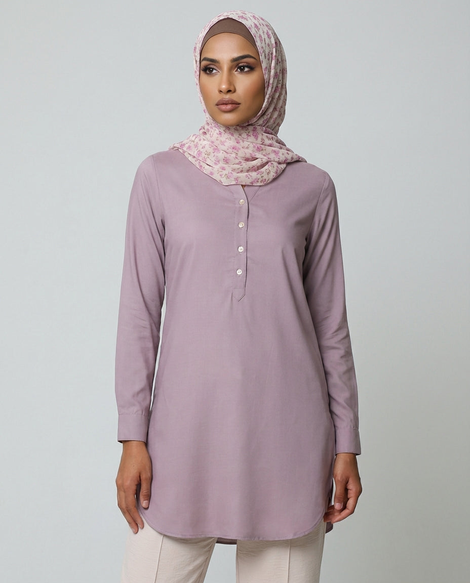 Woman wearing a light purple long-sleeve top and white pants with a patterned hijab on a gray background