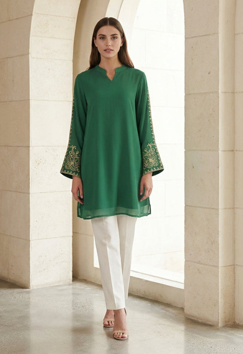 Woman wearing a green embroidered top and white pants in an arched hallway.