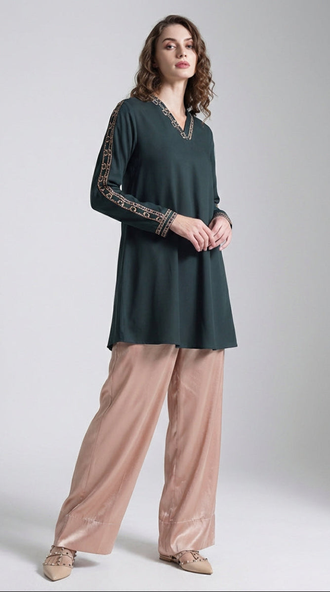 Woman wearing a dark green top with embroidered sleeves and beige pants on a white background
