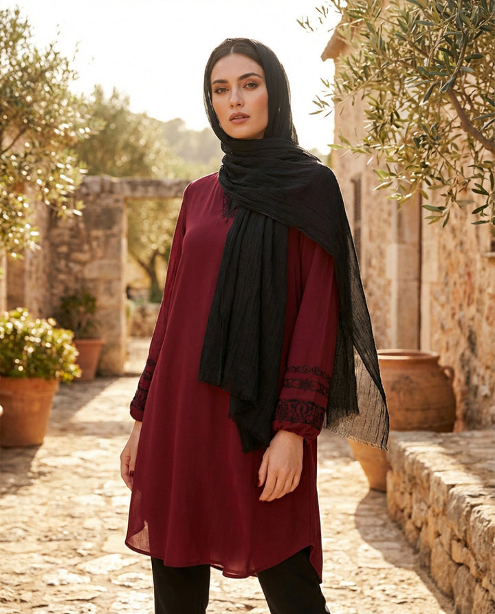Woman wearing a red dress and black hijab standing outdoors with stone walls and potted plants in the background.