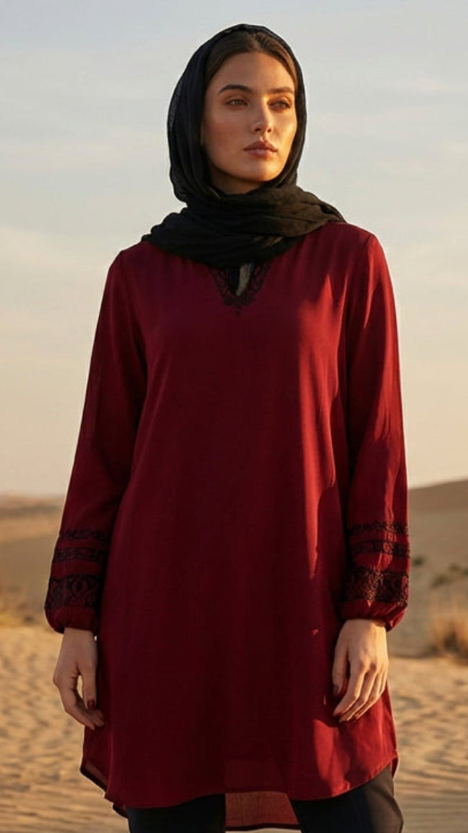 Woman in a red dress and black hijab standing in a desert landscape with sand dunes and sparse vegetation.