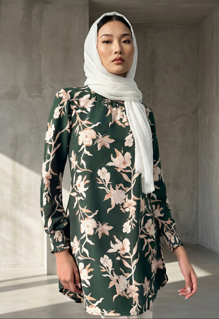 Woman wearing a floral dress and white hijab in a sunlit room.