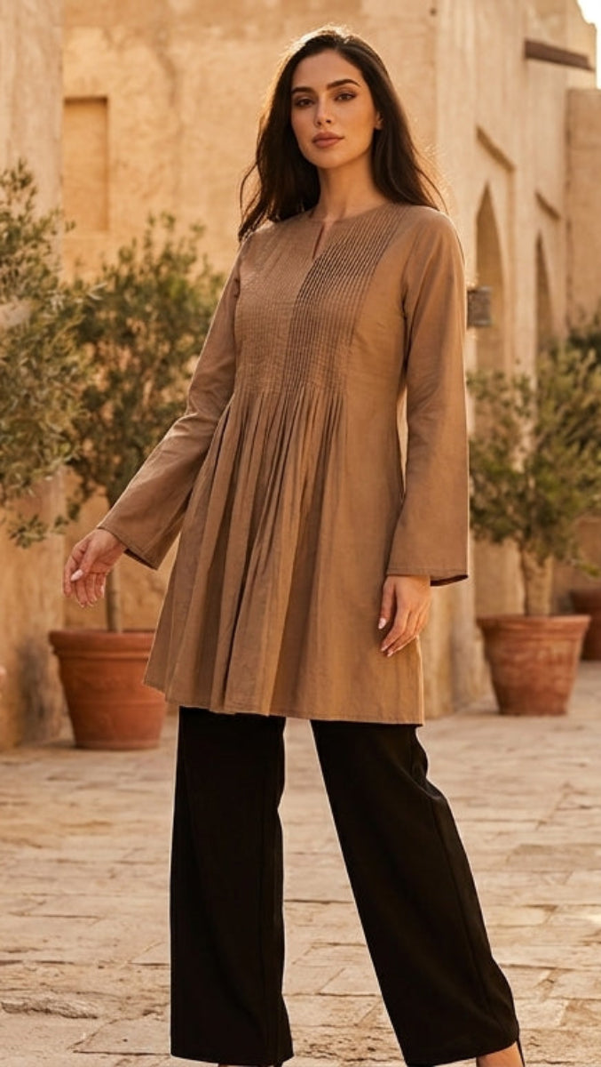 Woman in a beige long-sleeve top and black pants standing in a sunlit courtyard with stone walls and potted plants.