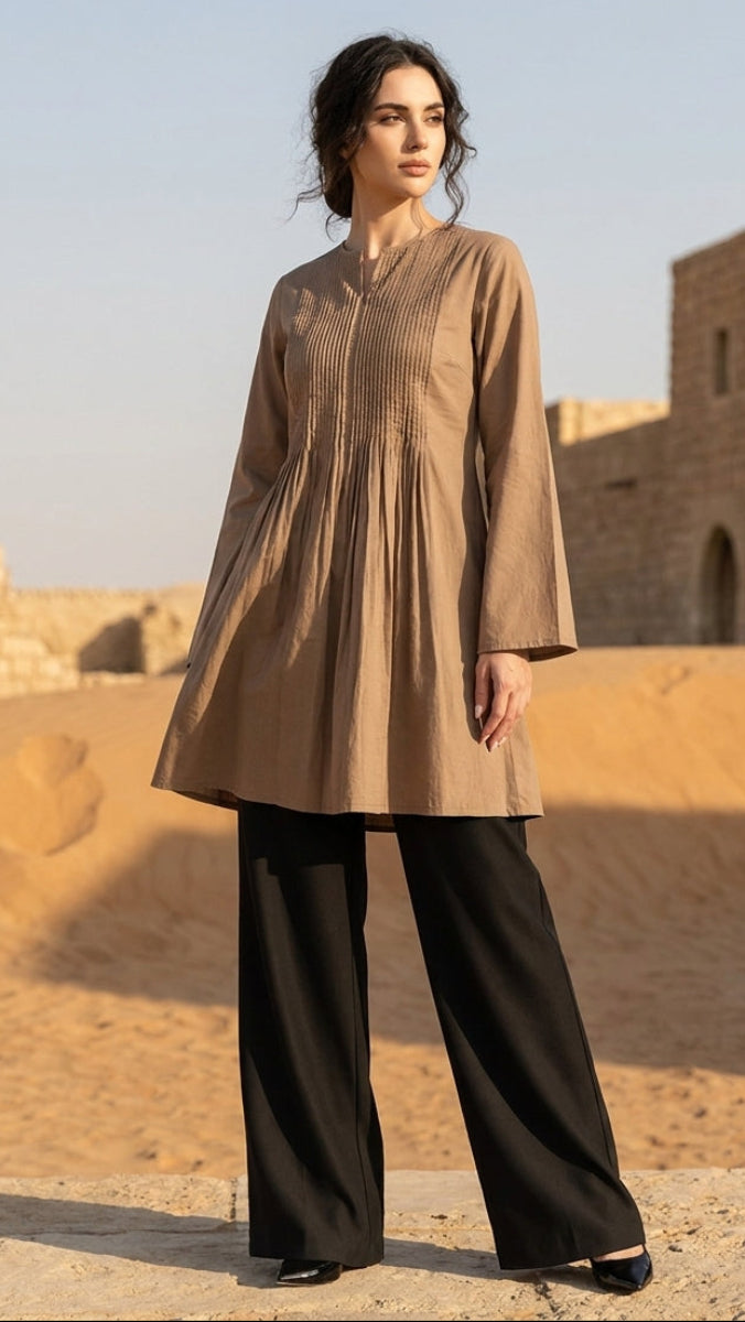 Woman in a beige long shirt and black pants standing in a desert with ancient architecture.