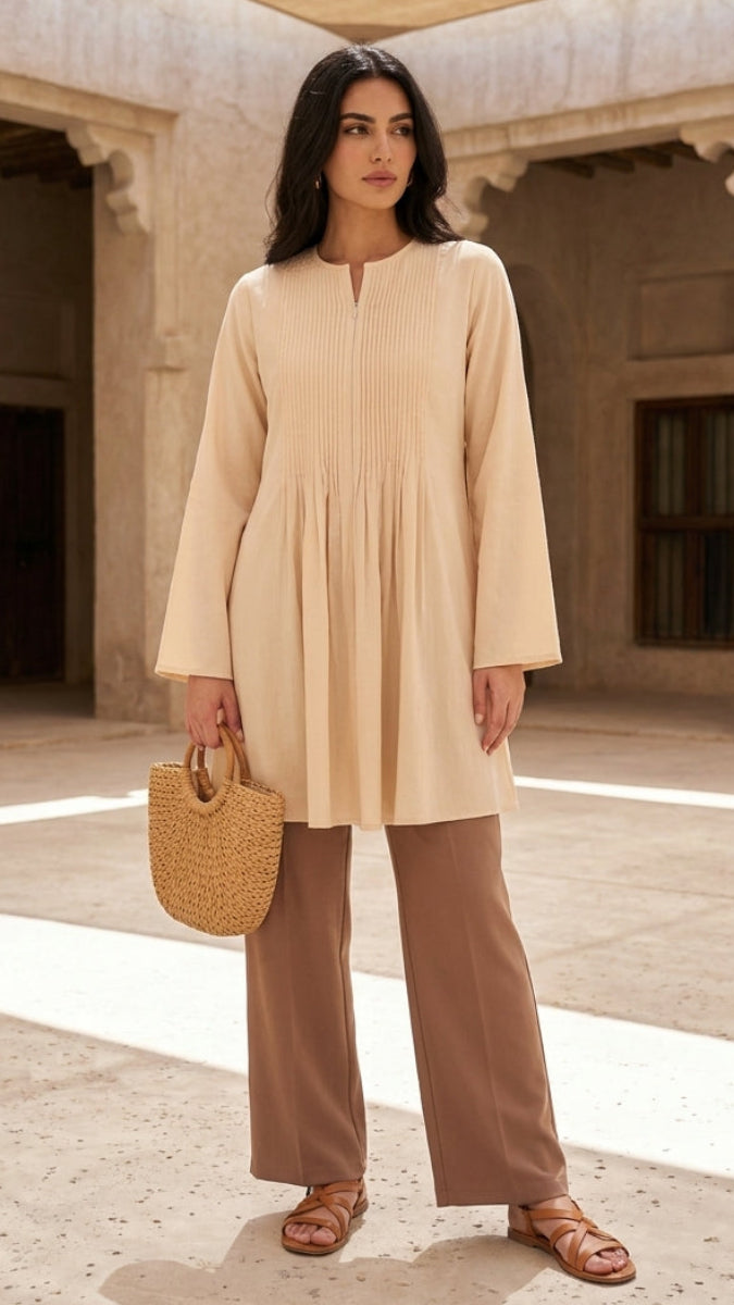 Woman in beige long-sleeve top and brown pants holding a straw bag in an architectural setting.