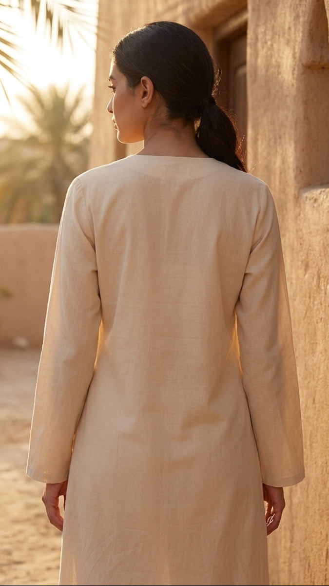 Woman in a beige dress standing in a desert setting with palm trees and sand dunes.