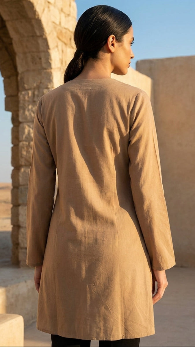 Woman wearing a beige coat standing in front of stone architecture with a clear blue sky.
