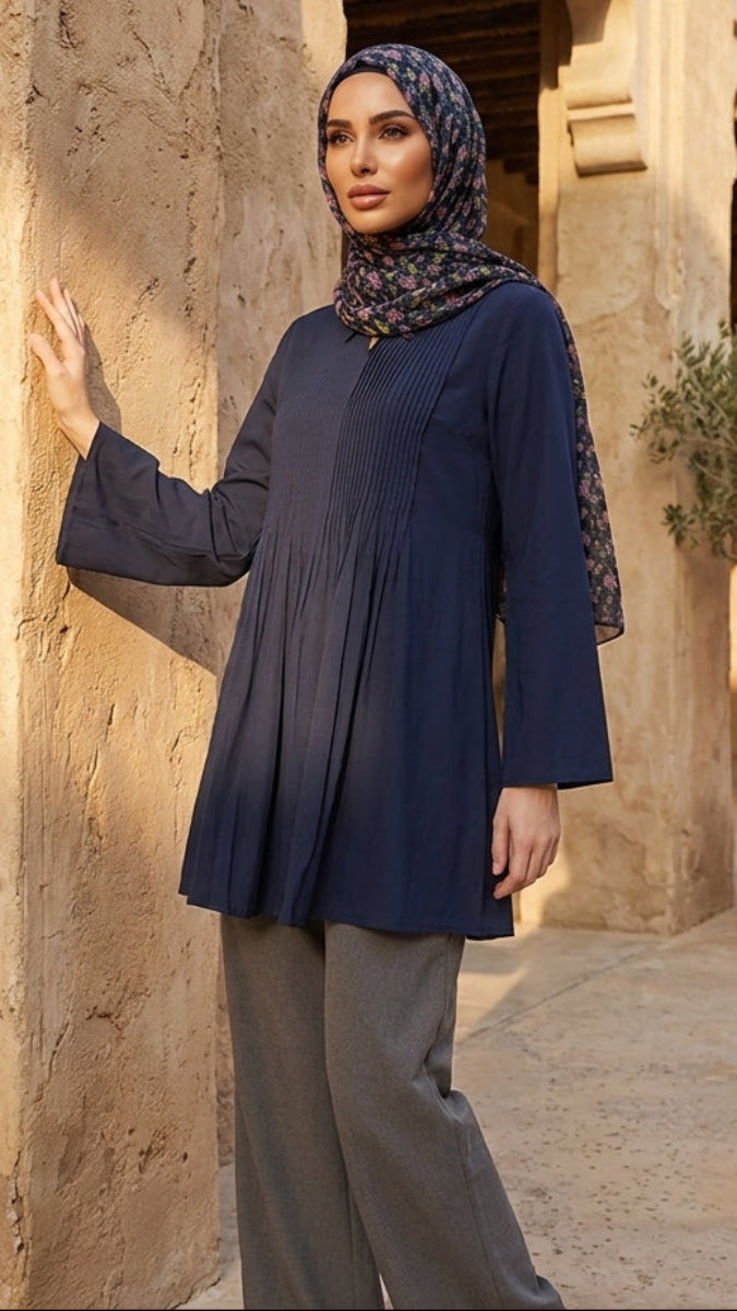Woman in a blue top and hijab standing in front of a stone building with potted plants.