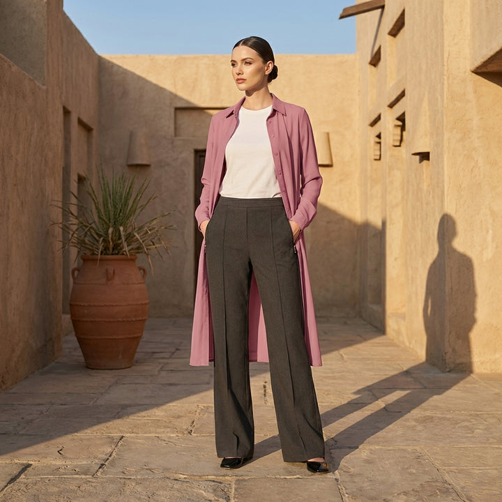 Woman in a pink coat and black pants standing in a sunlit courtyard with a plant and stone wall.