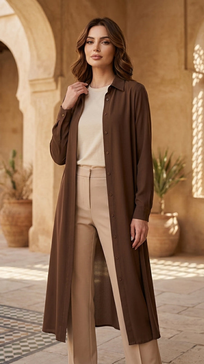 Woman wearing a long brown coat over a white top and beige pants in a sunlit courtyard.