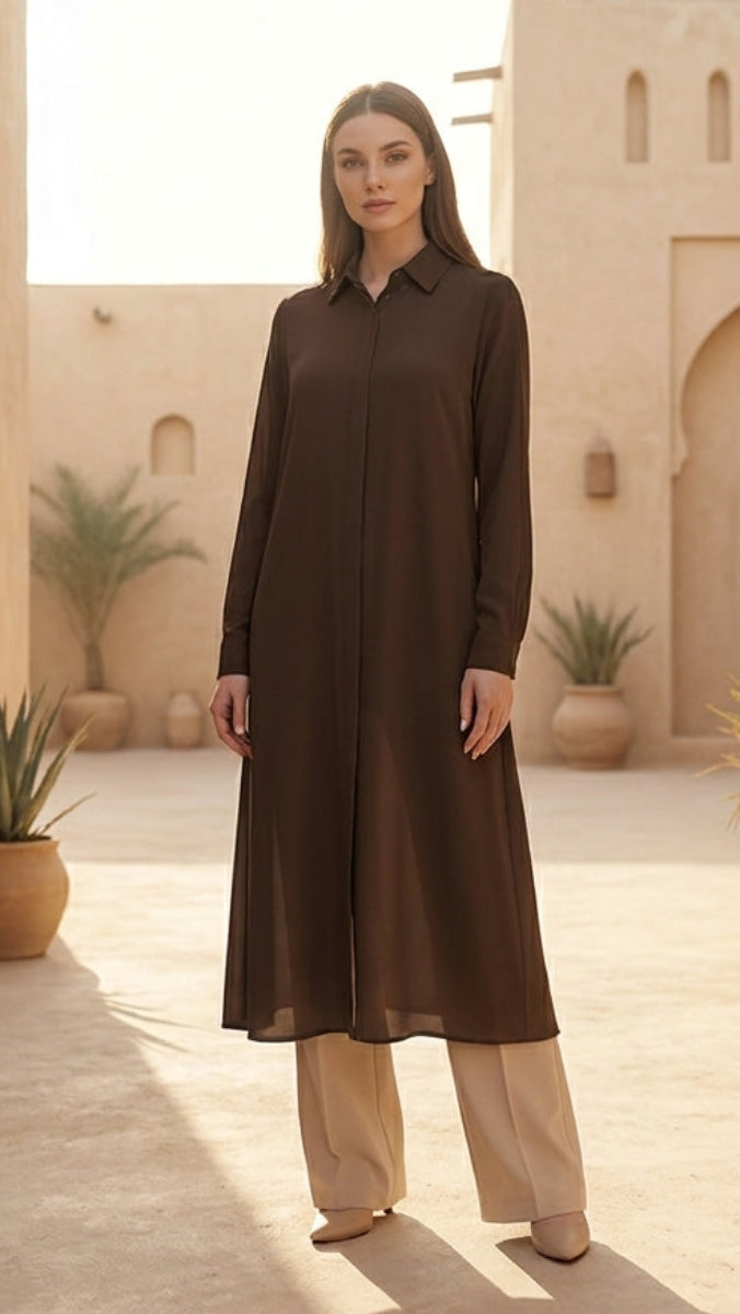 Woman in a long brown dress standing in a sunlit courtyard with architectural elements and plants.