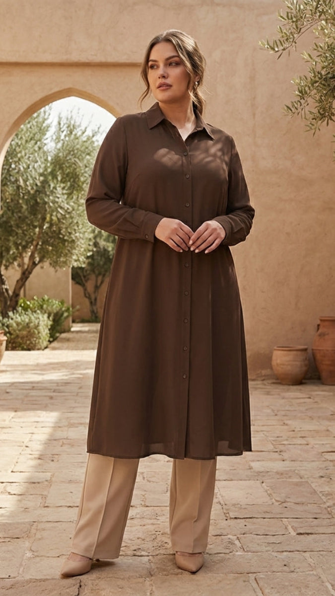 Woman in a brown dress standing in a sunlit outdoor setting with beige walls and potted plants.