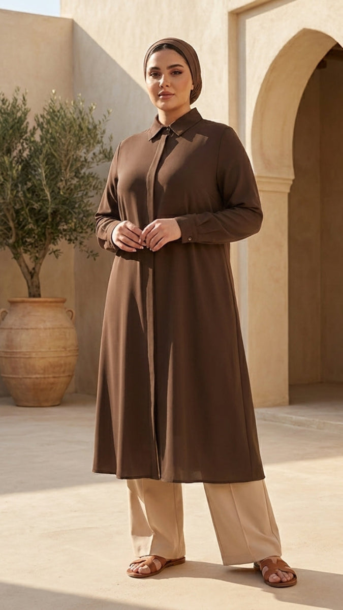 Woman in a long brown dress standing in a sunlit courtyard with arches and a potted plant.
