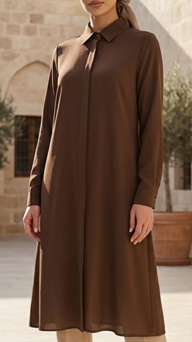 Woman in a brown outfit standing in a sunlit courtyard with stone architecture and potted plants.