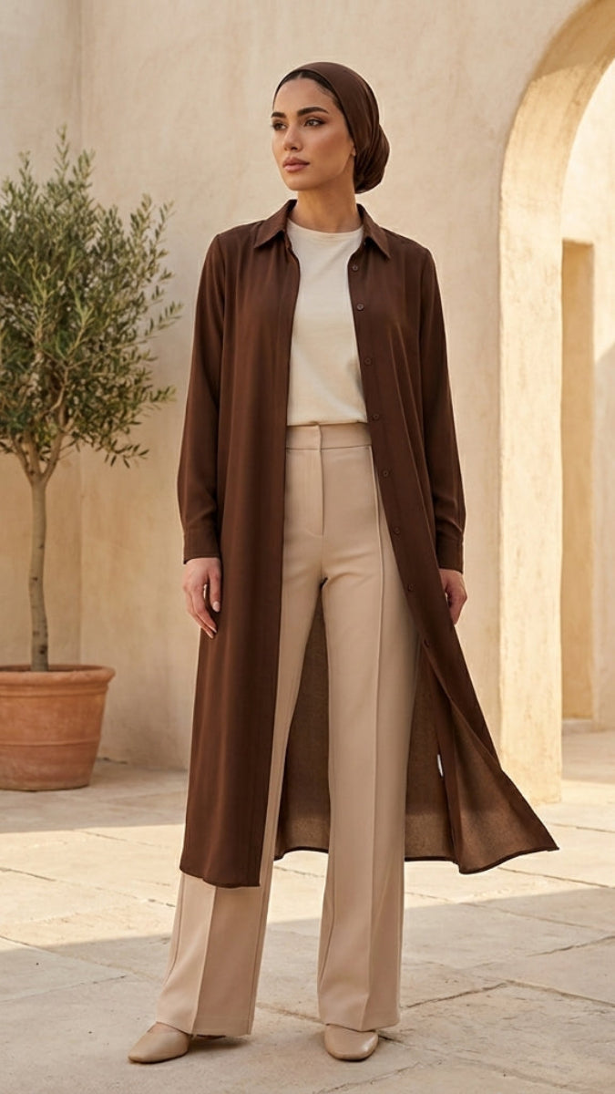 Woman in a brown coat and beige pants standing in a sunlit archway with a potted plant.