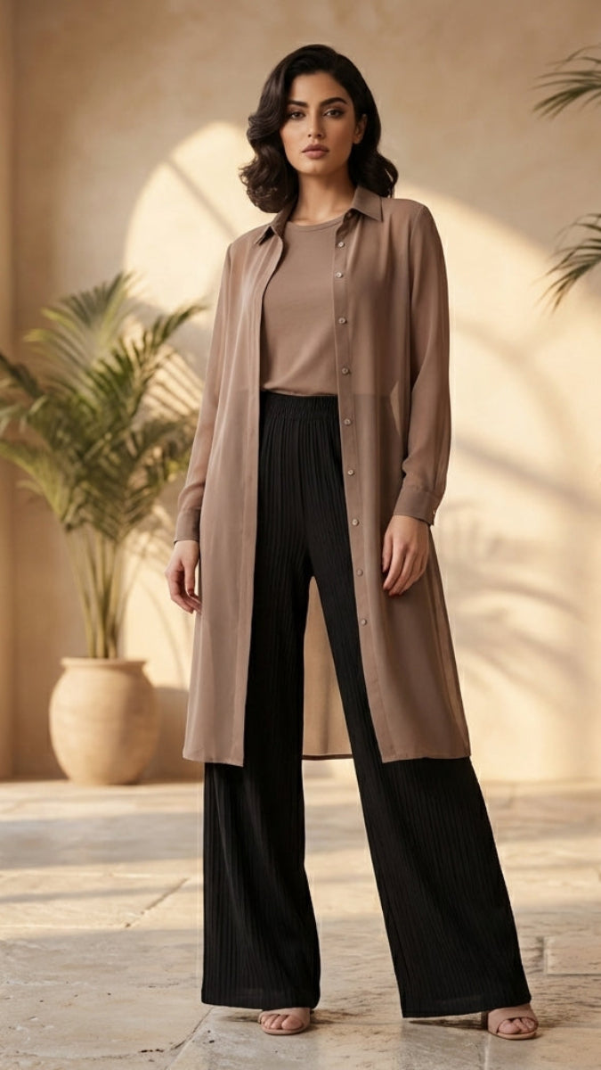 Woman in a beige long coat and black pants standing in a sunlit room with plants.