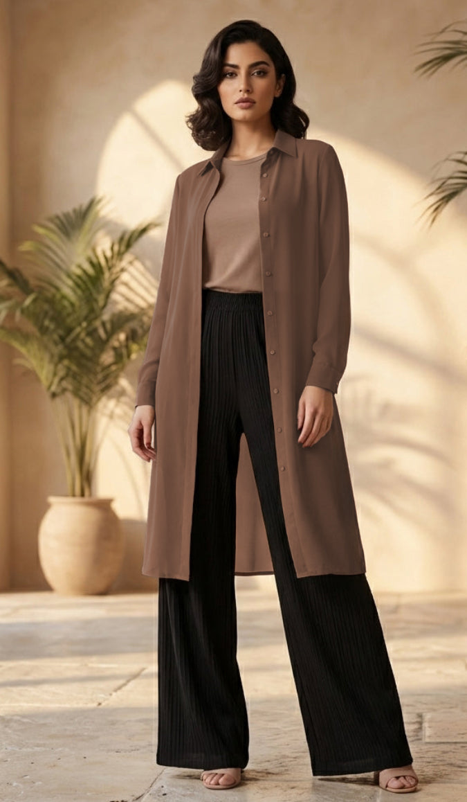 A woman models a long-sleeve, button-front brown cardigan over a beige top and black wide-leg pants in a warm, sunlit indoor setting with potted plants.