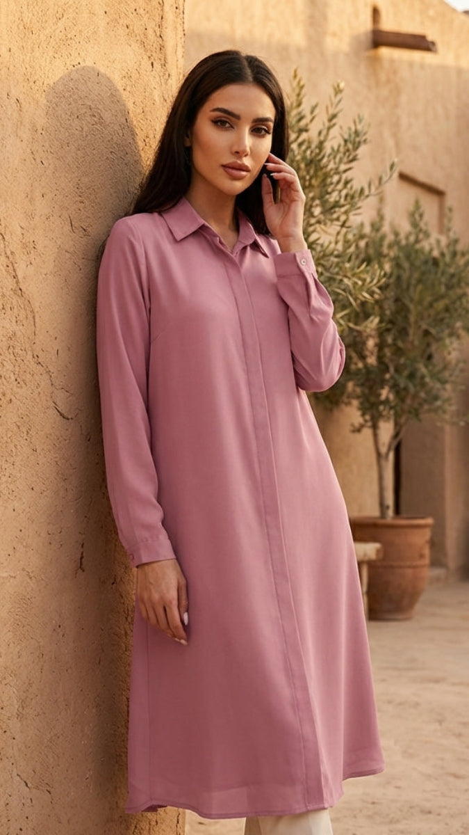Woman in a long pink dress standing against a textured wall with potted plants in the background.