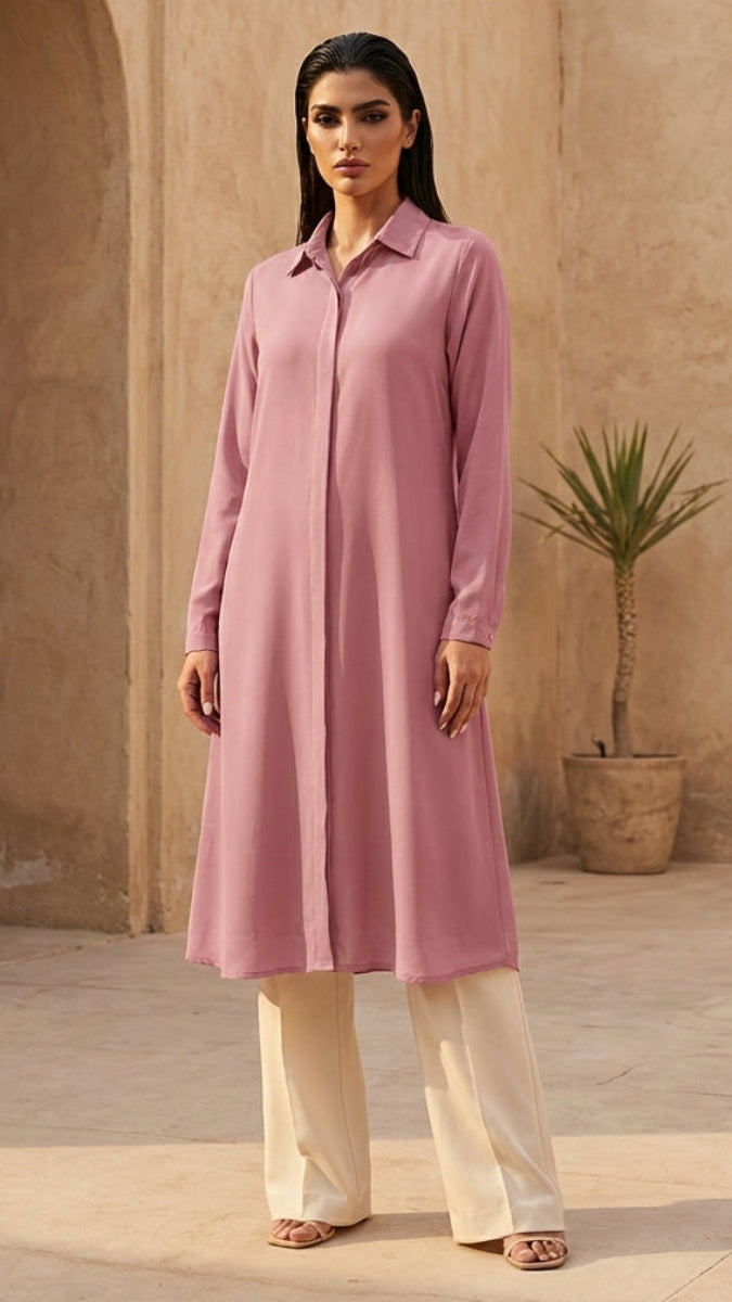 Woman wearing a pink long shirt and white pants standing in a sunlit courtyard with arches and a plant.