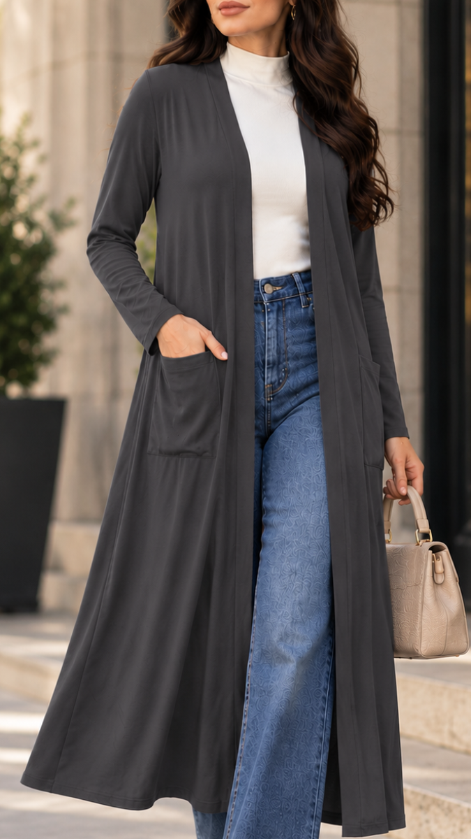 A woman wearing a long dark gray open-front cardigan over a white mock-neck top and blue jeans, holding a beige handbag in an outdoor setting.