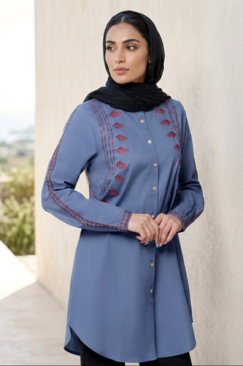 Woman wearing a blue embroidered dress with a black hijab outdoors.
