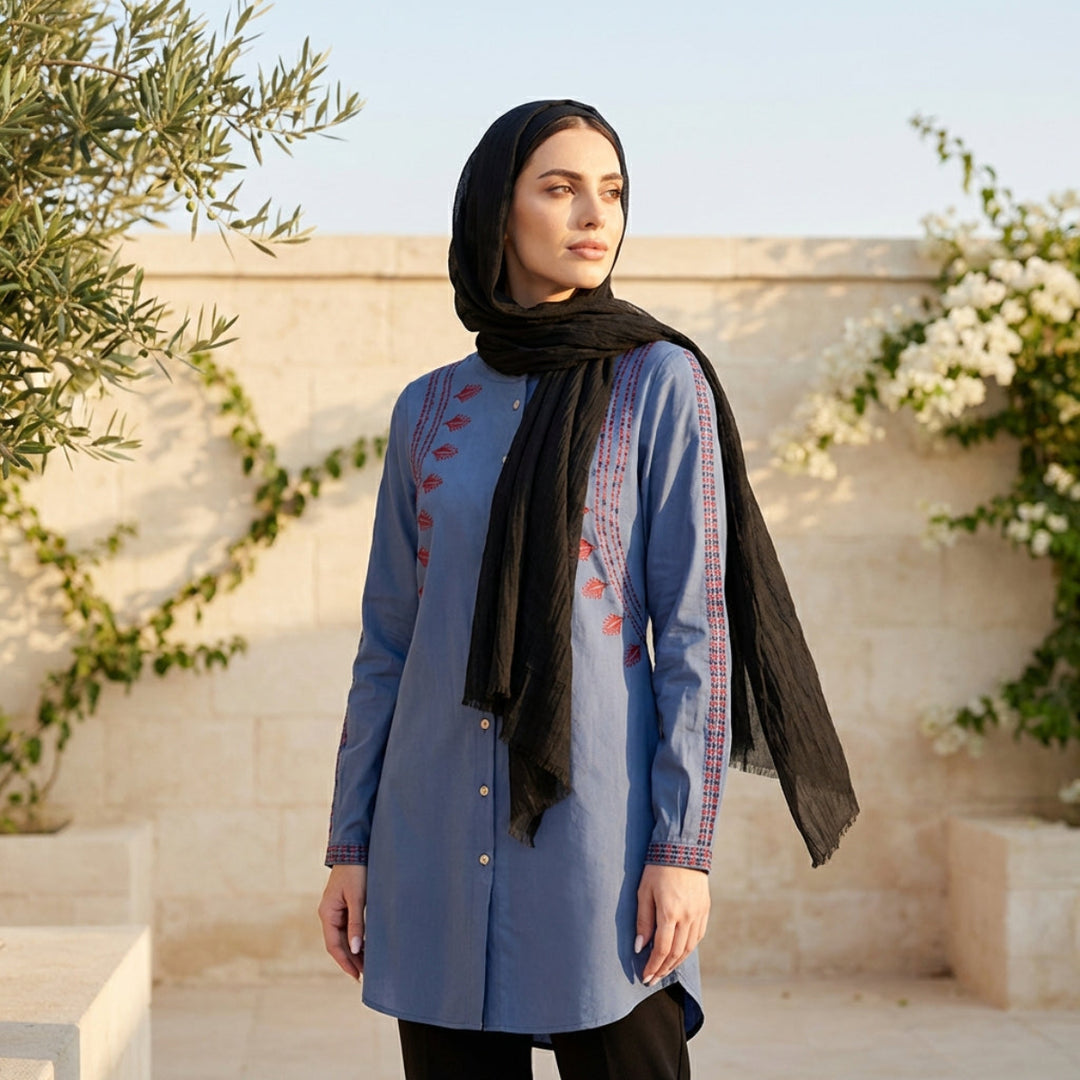 Woman wearing a blue embroidered dress with a black hijab in an outdoor setting.