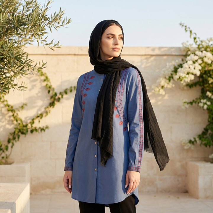 Woman wearing a blue embroidered dress with a black hijab in an outdoor setting.
