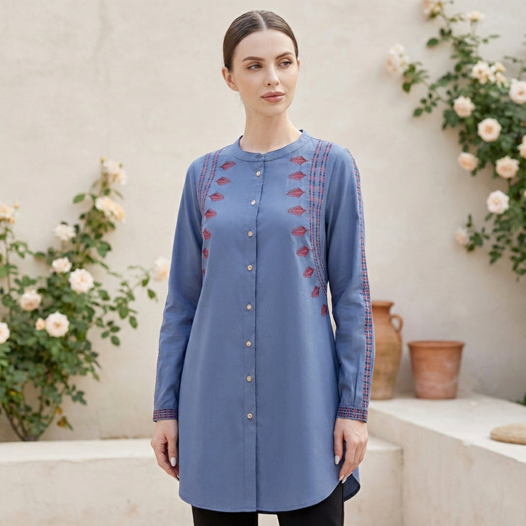 Woman wearing a blue embroidered tunic standing in front of floral decorations.