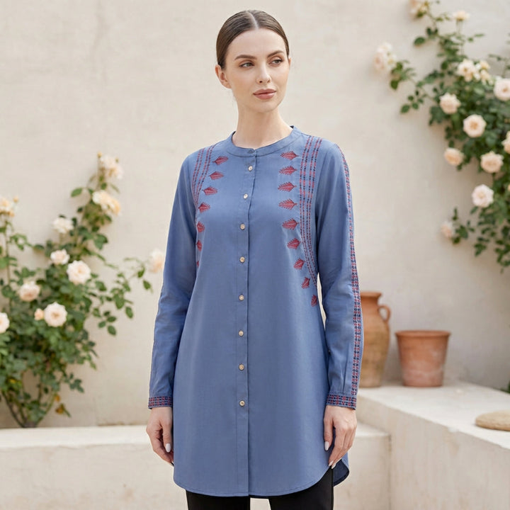 Woman wearing a blue embroidered tunic standing in front of floral decorations.