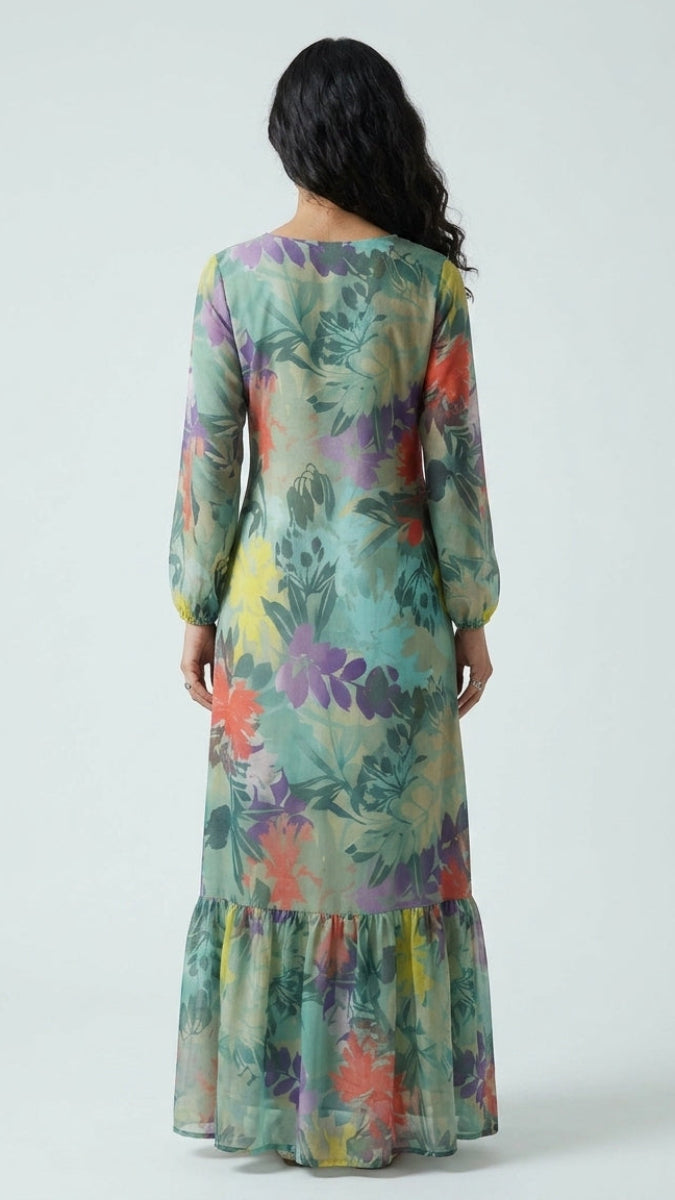 A woman viewed from the back wearing a long-sleeve maxi dress with a colorful floral and botanical print on a light teal-green base, set against a plain light gray studio background.