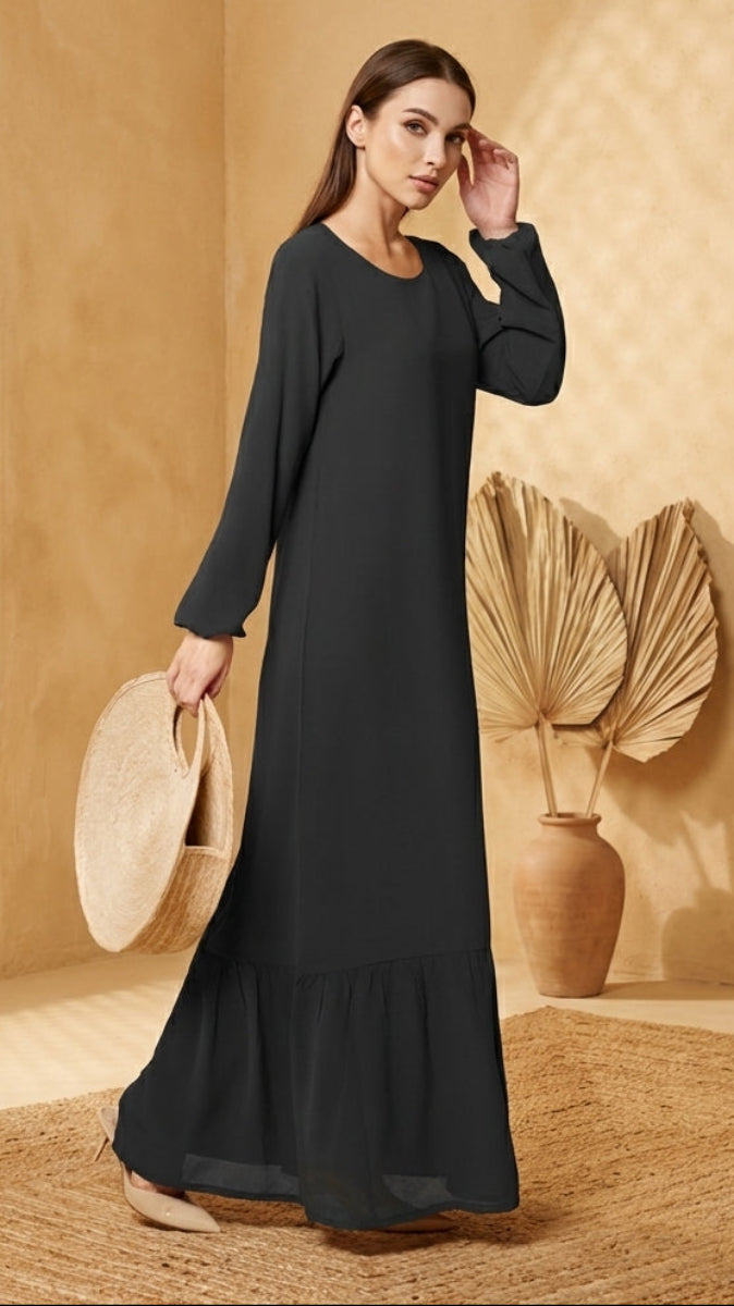 Woman in a long black dress holding a hat in a warm-toned room.