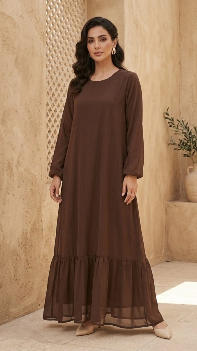 A woman modeling a long-sleeved, tiered brown maxi dress in a warm, sandy-colored architectural setting.