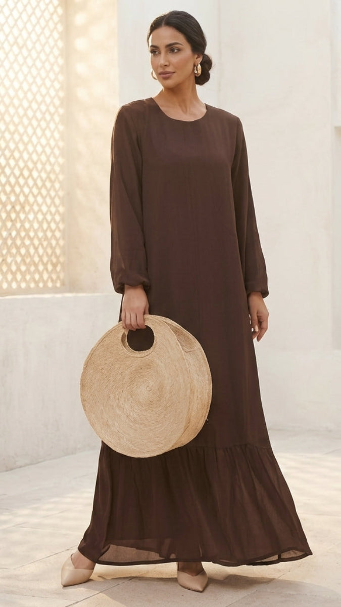 A woman wearing a long-sleeved, dark brown maxi dress and holding a round woven straw bag, standing in a bright, sunlit architectural setting.