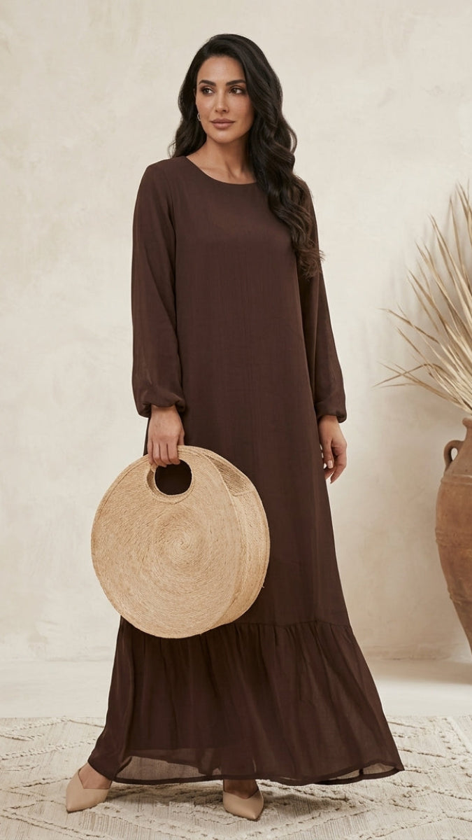 A woman modeling a long-sleeved, dark brown tiered maxi dress, holding a round woven straw tote bag against a neutral beige textured wall background.