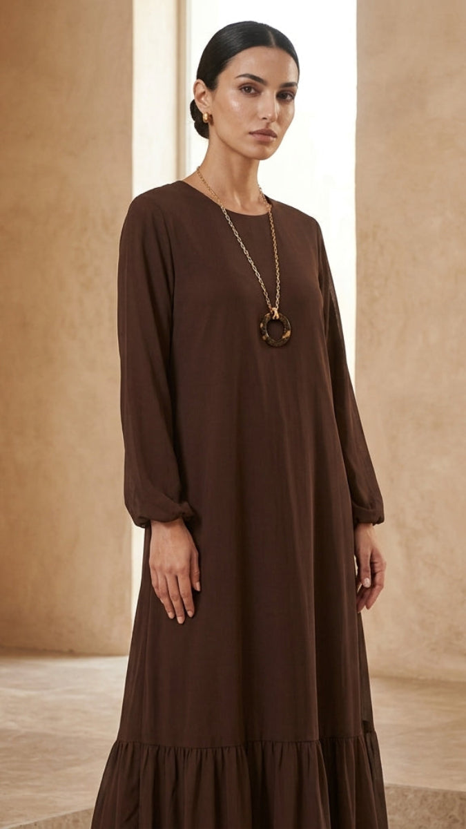 A woman modeling a long-sleeved brown maxi dress with a tiered hem, standing in a warm, neutral-toned architectural setting.