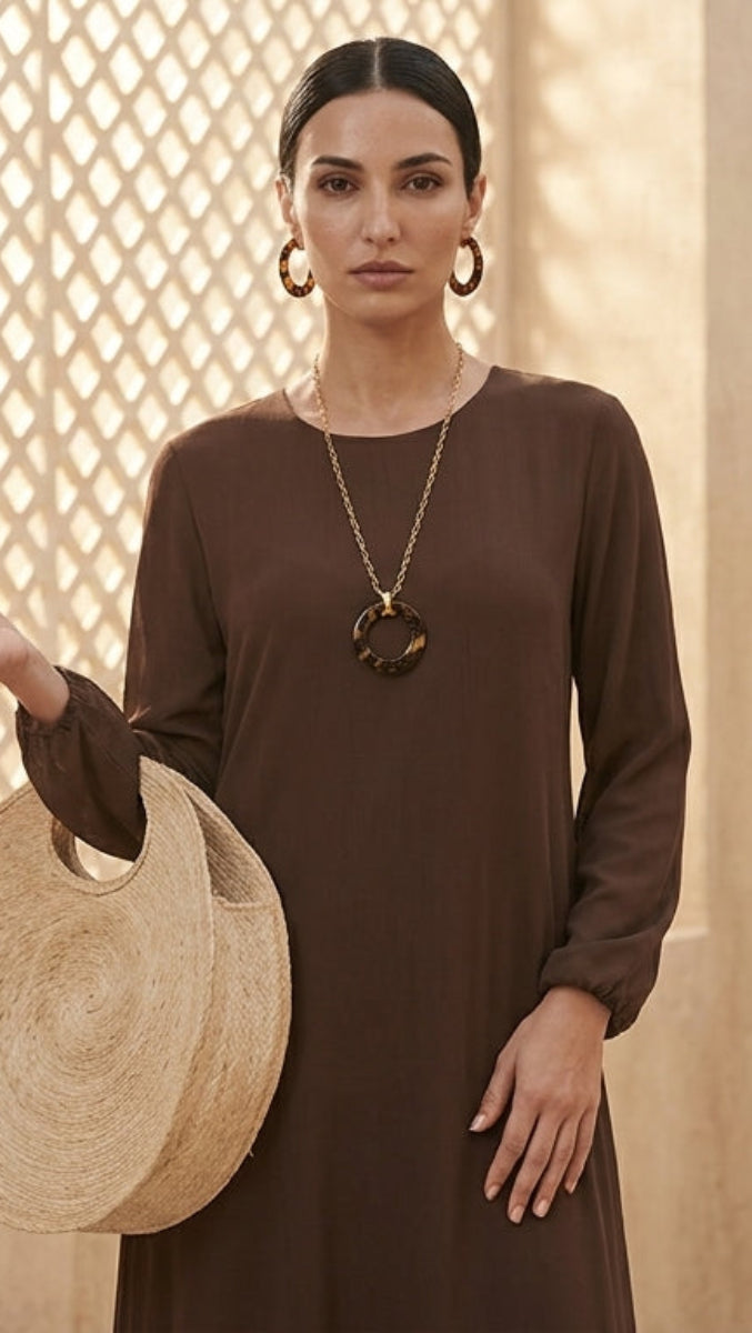 A woman wearing a dark brown long-sleeve dress and holding a round woven straw hat, standing in a warm-toned beige room with a lattice-patterned window.
