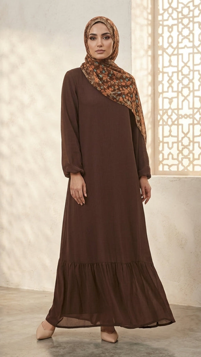 A woman wearing a long-sleeved brown modest dress with a tiered hem and a patterned brown and orange hijab, standing in a softly lit interior with a decorative window screen.