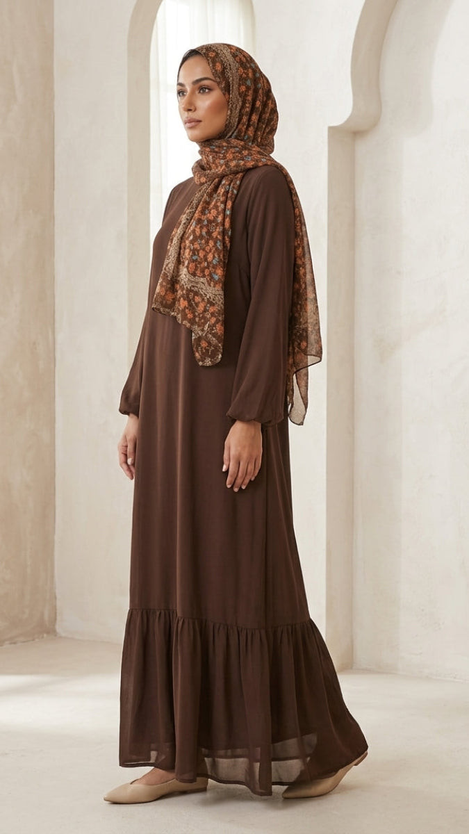 A woman wearing a long brown modest maxi dress with a tiered hem and a matching patterned brown hijab, standing in a bright, neutral-toned architectural setting.