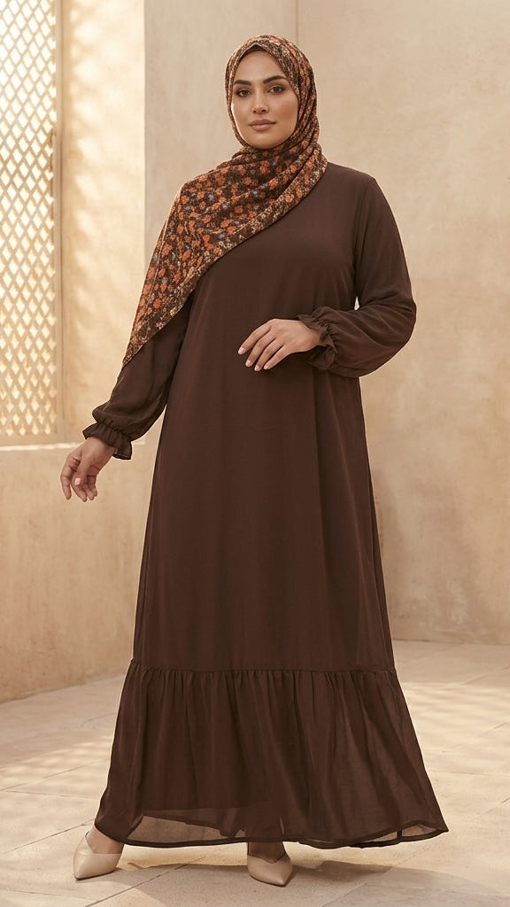 A woman wearing a long-sleeved brown abaya dress with a tiered hem and a matching brown patterned hijab, standing in a warm beige courtyard setting.