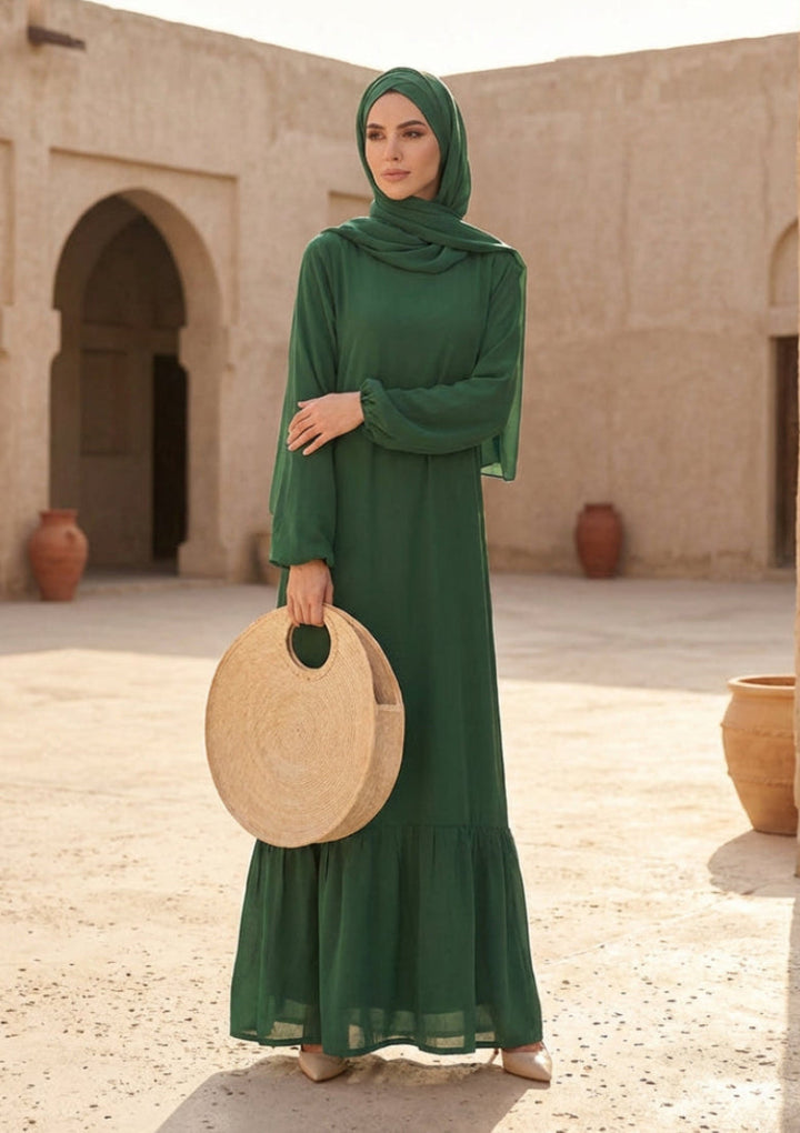 Woman in a green dress holding a round beige bag in an outdoor setting with beige walls and pots.