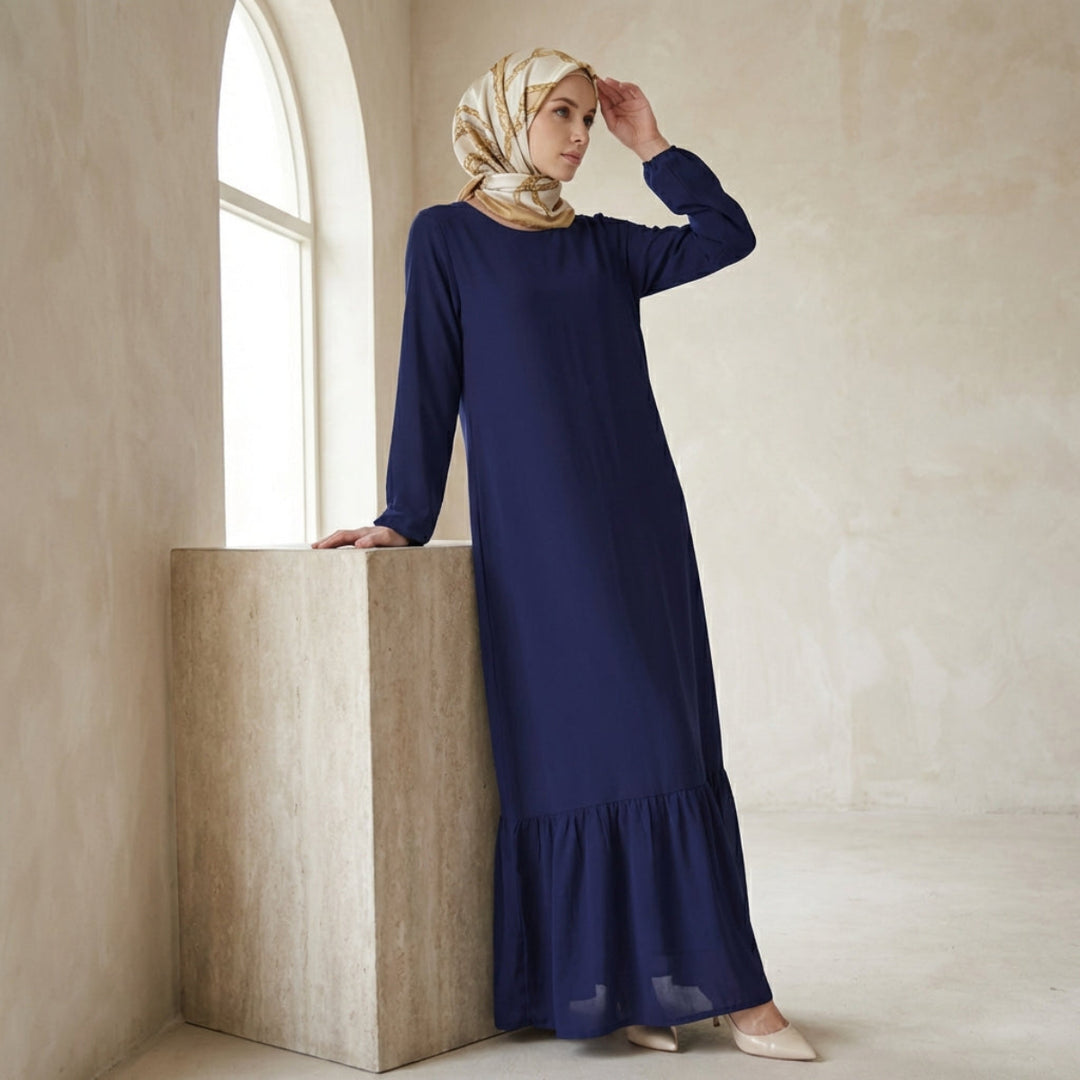 Woman in a long blue dress and headscarf standing in a softly lit room.
